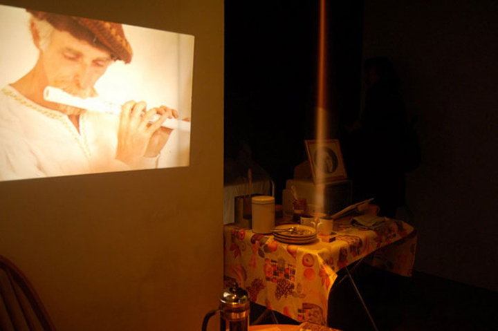 Installation still featuring sand pouring onto a table in a steady stream illuminated by the light of a beamer projecting the image of a man playing the flute. Coffee and cake are featured on a table in the background.