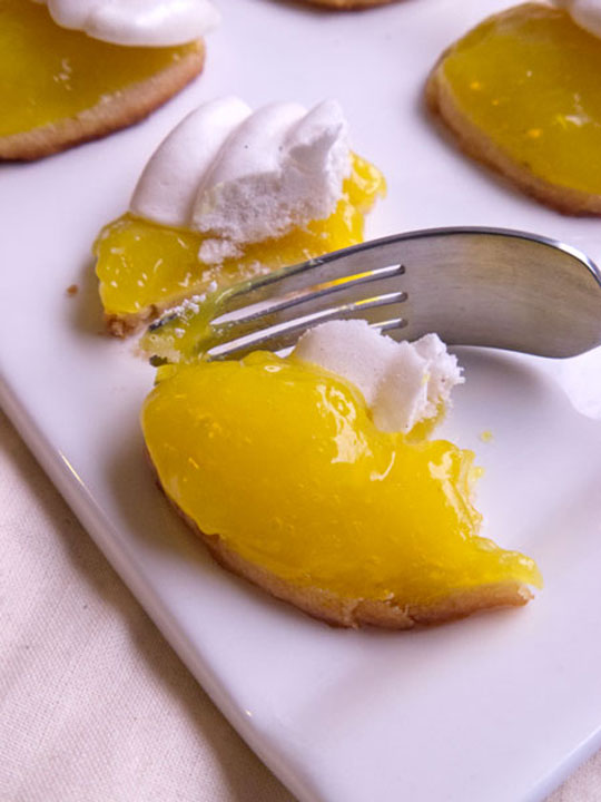 Picture of a small cookie topped with lemon curd and a squiggly shaped bit of meringue. The small pastry is cut in half with a fork and sits on a white plate with three other bites visible in the background.