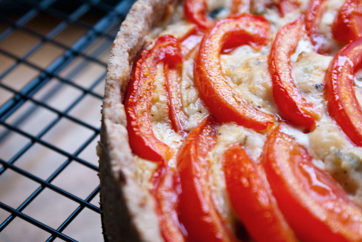 Close-up picture of a quiche with tomato slices arranges in a circular pattern. The quiche sits atop a cooing rack with small black metal squares that contrast with the table underneath it.