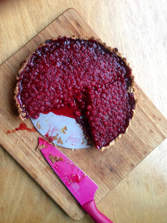 Picture of a redcurrant pie on a wooden cutting board on top of a wooden background. One-fifth has been cut out with a magenta knife also pictured.