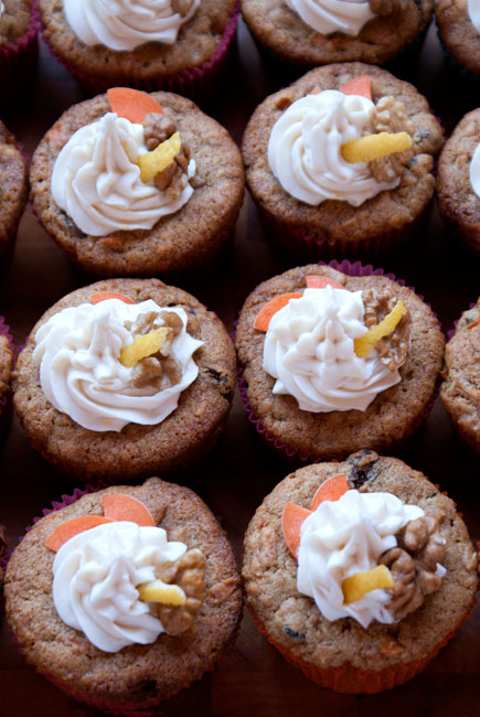 Picture of six carrot cupcakes with frosting and decorative slices of carrot, walnut and lemon zest as decoration.