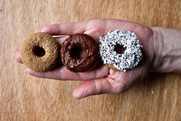 Picture of three miniature donuts, one with coconut topping, one with cinnamon sugar, one with chocolate frosting, placed on a horizontal line on a hand over a wooden tabletop.